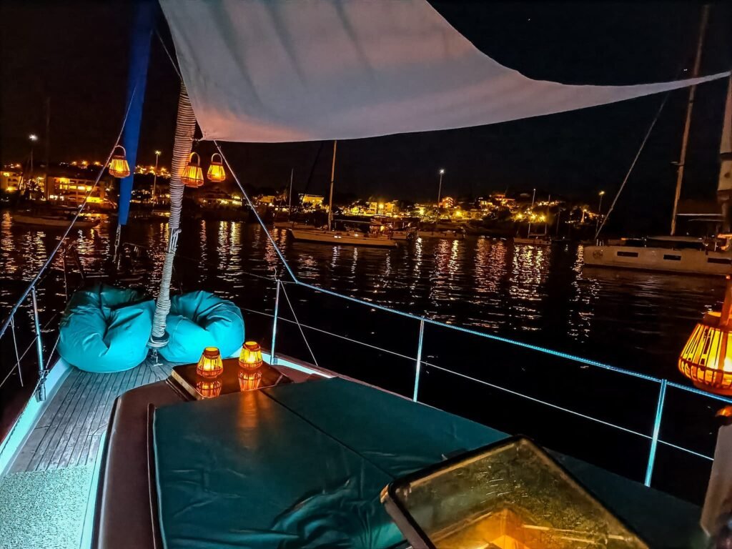 Proa de un velero de noche, con cojines y las luces de un puerto al fondo