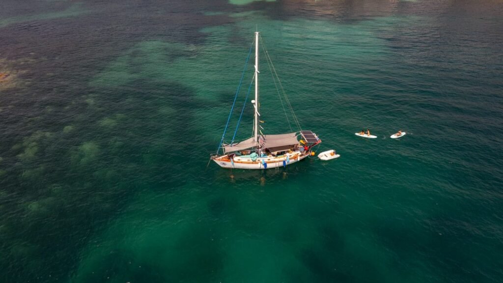 Vista aérea de un velero fondeado en aguas verdes, con una tabla de paddle surf cerca
