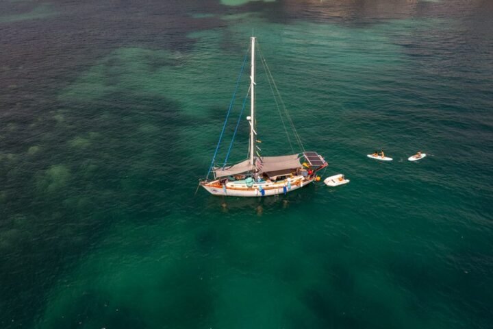 Vista aérea de un velero fondeado en aguas verdes, con una tabla de paddle surf cerca